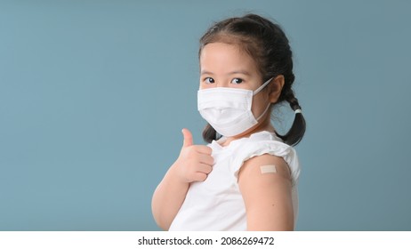 Coronavirus Vaccination Advertisement. Happy Vaccinated Little asian girl Showing Arm With Plaster Bandage After Covid-19 Vaccine Injection Posing Over Blue Background, Smiling To Camera. New normal.