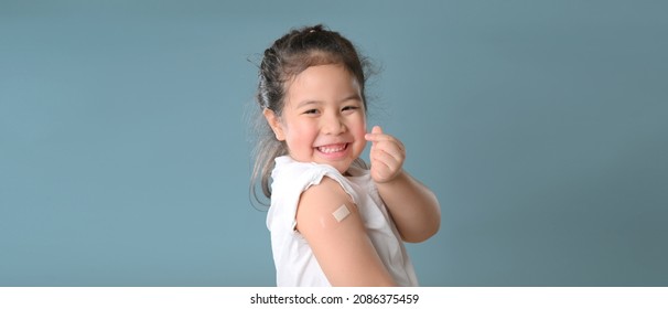 Werbung für die Impfung mit Coronavirus. Happy geimpftes kleines asiatisches Mädchen, das nach der Covid-19-Impfung auf blauem Hintergrund einen Arm mit Plaster-Bandage zeigt und dabei an die Kamera lächelt.  Leerraum