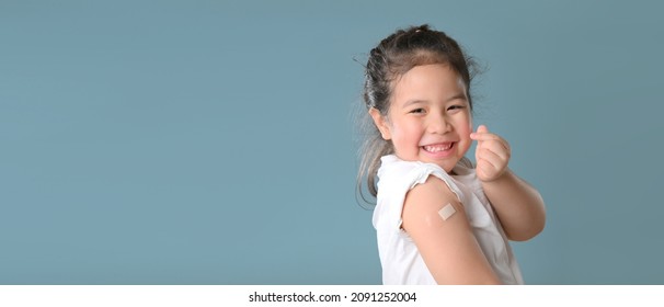 Werbung für die Impfung mit Coronavirus. Happy geimpftes kleines asiatisches Mädchen, das nach der Covid-19-Impfung auf blauem Hintergrund einen Arm mit Plaster-Bandage zeigt und dabei an die Kamera lächelt. Neu normal.