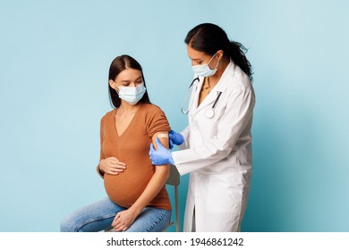 Coronavirus Vaccination And Pregnancy. Nurse Vaccinating Pregnant Female Patient Against Covid-19 Sticking Bandage After Vaccine Injection In Arm Over Blue Studio Background, Wearing Face Masks