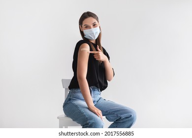 Coronavirus vaccination saves lives. Young woman in face mask pointing at adhesive bandage on her arm, getting vaccinated against covid-19 on light background. Immunization for infectious disease