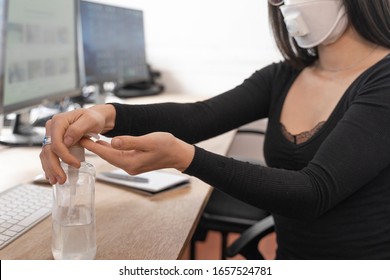Coronavirus. Woman cleaning her hands at the office. Sick with mask for corona virus. Workplace desk with computer. Woman spraying alcohol gel or antibacterial soap sanitizer.