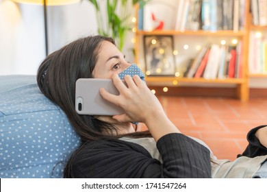 Coronavirus. Woman lying down using mobile phone. Wearing mask protection and recovery from the illness in home. Quarantine. Patient isolated to prevent infection. Pandemia. Home.