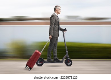 Corporate businessman riding an eco-friendly electric scooter and carrying a roller suitcase, city in the background, sustainable mobility concept - Shutterstock ID 2228871123