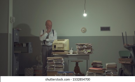 Corporate Businessman Searching For Files In A Rundown Messy Office With An Old Computer And Cheap Furniture, And Piles Of Paperwork