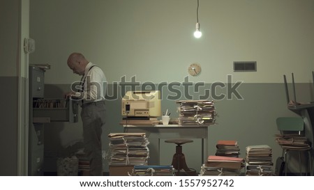 Corporate businessman searching for files in a rundown messy office with outdated computer and piles of paperwork
