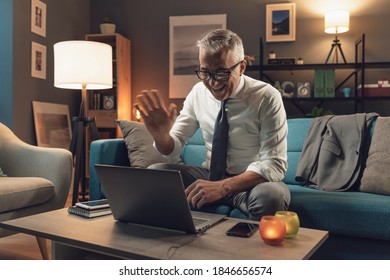 Corporate Businessman Sitting On The Sofa At Home And Connecting With His Laptop, He Is Videocalling Someone And Waving At The Computer Webcam