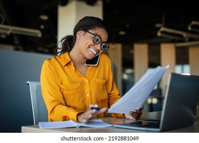 Corporate communication. Happy black female manager working with papers and talking on mobile phone, reading financial documents while sitting at workplace in office - Powered by Shutterstock