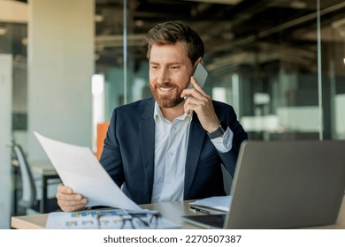 Corporate communication. Happy male manager working remotely, reading financial document and talking on mobile phone, sitting at desk in office interior - Powered by Shutterstock