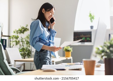 Corporate Communication. Portrait of smiling young Asian female manager working remotely, holding paper and reading financial document, talking on mobile phone standing at desk at home office - Powered by Shutterstock