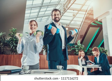 Corporate Lifestyle Healthcare Concept Of Business People In Office Interior Working With Dumbbells. Workers Doing Fitness Exercises Of Yoga Practice. Wellbeing Employee Man Smiling To Healthy Woman