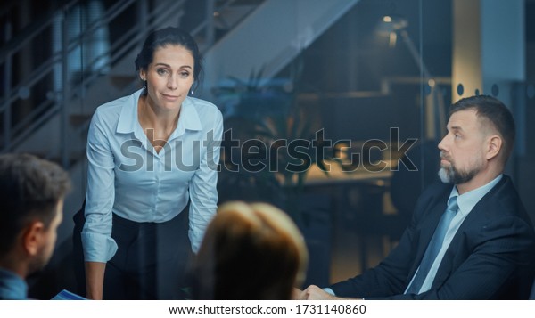 Corporate Meeting Room: Confident and Beautiful\
Female Executive Director Decisively Leans on the Conference Table\
and Delivers Report to a Board of Executives about Company’s Record\
Breaking Revenue