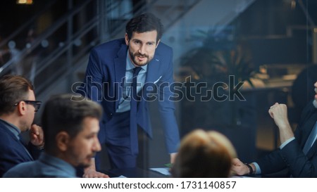 Corporate Meeting Room: Confident and Handsom Executive Director Decisively Leans on a Conference Table and Delivers Report to a Board of Executives about Company’s Record Breaking Revenue
