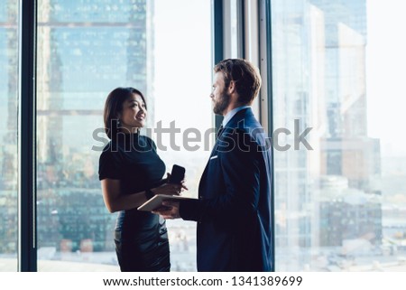 Corporate multicultural employees standing near window in office interior and talking about work, two successful cheerful business colleagues conversating and sharing ideas. Workers discussing ideas