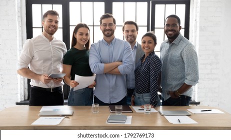 Corporate photo smiling diverse employees with confident executive wearing glasses standing in modern office room, looking at camera, successful startup founder with team, staff members