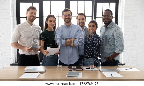 Corporate photo smiling diverse employees with\
confident executive wearing glasses standing in modern office room,\
looking at camera, successful startup founder with team, staff\
members