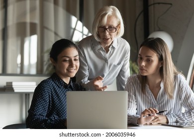 Corporate teacher showing and explaining work data to student girls, pointing at laptop screen, speaking. Mentor training interns. Elder coach giving advice to diverse office apprentices - Powered by Shutterstock