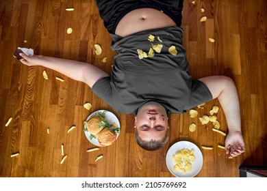 Corpulent Fat Boy Eat Junk Food While Lying On Floor At Home, With French Fries And Chips. Top View Portrait Of Obese Teenager Boy In Casual Wear Spending Time Alone, Lead Unhealthy Lifestyle