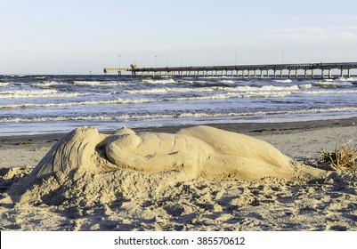 CORPUS CHRISTI, TEXAS/USA - FEBRUARY 26, 2016: A Sand Sculpture Of A Mermaid By An Unidentified Artist Graces The Beach Near Sunset At Padre Balli Park On Padre Island, With Bob Hall Pier Beyond.