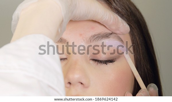 Correction of a shape of eyebrows with hot wax. Brow
master applying wax on the eyebrow of female face. Wax correction
of the shape of the eyebrows with spatula. Beauty industry. Close
up. 