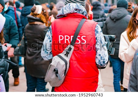 Correspondent in a red jacket with text “Press” and camera working in crowd. Protest. Strike. City. Outdoor. Job. Professional. Occupation. Specialist. News. Media. Broadcast. TV