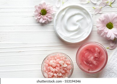 Cosmetic Cream Container, Body Scrub, Bath Salt And Pink Flowers On White Wooden Background From Top View