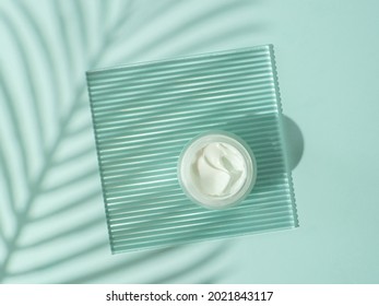 Cosmetic cream or moustirizer on transparent ribbed acrylic plate with tropical palm leaf shadows on blue background. Open round glass jar with aesthetic swirls cream. Top view flatlay. Copy space
