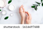 Cosmetic cream on female hands, jars with milk swirl cream and green leaves on white marble table. Flat lay, top view. Woman applying organic moisturizing hand cream. Hand skin care concept