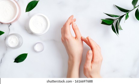 Cosmetic cream on female hands, jars with milk swirl cream and green leaves on white marble table. Flat lay, top view. Woman applying organic moisturizing hand cream. Hand skin care concept - Shutterstock ID 1616697688