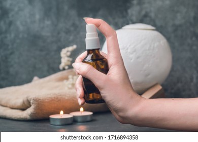 Cosmetic dark amber glass bottles in female hands on dark gray background. Closeup, copyspace. Dark glass containers with spa candles and flowers. Perfume spray, salon treatment concept, minimalism