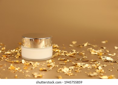 Cosmetic Glass Jar With White Moisturizer. Luxurious Anti-aging Day Cream With Isoflavones And Active Microparticles Of Gold And Pieces Of Gold Paper On Golden Background. Front View, Mockup.  