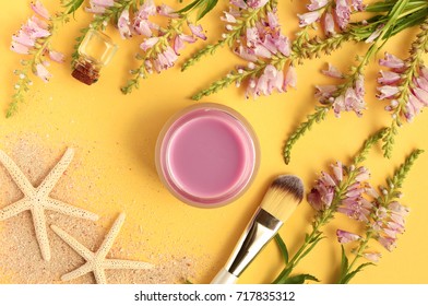 Cosmetic Herbal essence. Botanical summer skincare. Violet flowers, facial mask in jar with application brush, top view bright yellow background, starfish and sand 