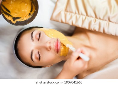 Cosmetic jelly mask, facial skincare. Cosmetologist applies yellow gold alginate mask with spatula on face of a woman. Facial skin treatment in the beauty salon. Top View above