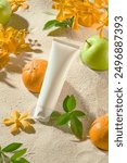 Cosmetic mock up was taken from above view on the white sand flat. In the middle, featuring a white unlabeled cosmetic tube, which leaning on an orange, apple, green plant and flower placed around