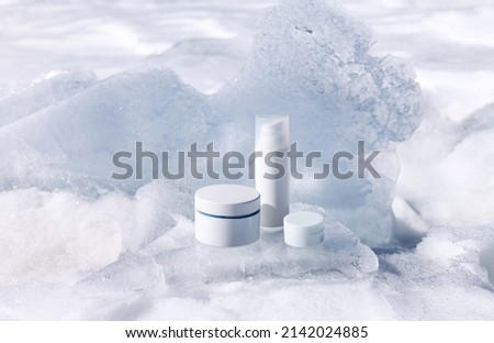 Cosmetic products for face and hand skin care in winter time. Advertising of nourishing cosmetics for cold weather on podium stand from piece of ice against backdrop of snow. White plastic packagings