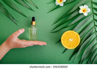 Cosmetic serum Vitamin C in glass bottle with pipette dropper in female hand. Orange essential oil with citrus Vitamin C and palm leaves on color green background. Natural skin care Cosmetics.