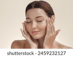 Cosmetics skincare concept photo. Woman with beautiful face touching healthy facial skin portrait. Beautiful smiling Asian girl model with natural makeup enjoys glowing hydrated smooth skin on beige