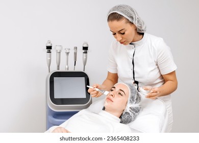 A cosmetologist applies anesthetic cream to a young woman's face before a facial cosmetic procedure. Anesthetic cream. Preparation for healing, tightening, skin rejuvenation in beauty salons. - Shutterstock ID 2365408233