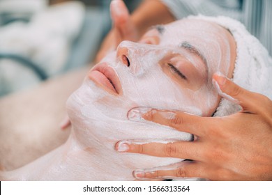 Cosmetologist applying rejuvenating facial mask onto woman’s face in beauty salon. 