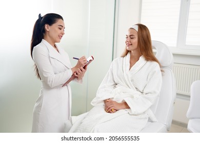 Cosmetologist consulting female patient and making notes during medical appointment in modern beauty clinic