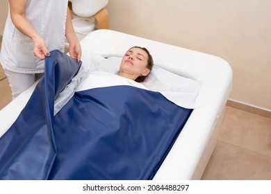 Cosmetologist covers the girl with a special blanket in the hydromassage bath. The procedure is a wrap in combination with a non-contact hot tub. Body and skin care concept. Cosmetology.