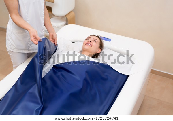Cosmetologist
covers the girl with a special blanket in the hydromassage bath.
The procedure is a wrap in combination with a non-contact hot tub.
Body and skin care concept.
Cosmetology.