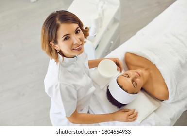 Cosmetologist or dermatologist looking up at camera near realxing woman in beauty salon