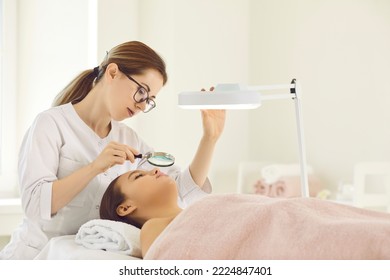 Cosmetologist examine female patient skin with magnifying glass do beauty procedures in aesthetic medicine clinic. Beautician or dermatologist do skincare treatment for woman in saloon. Cosmetology. - Shutterstock ID 2224847401