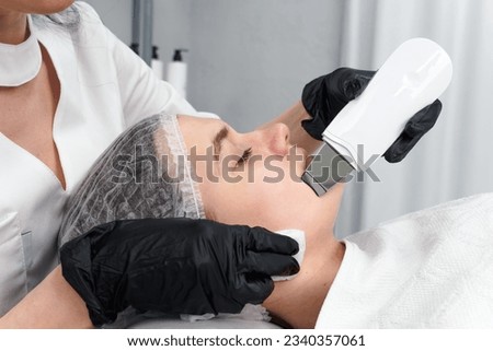 Cosmetologist making facial treatment with ultrasonic spatula to young woman. Ultrasound facial peeling at cosmetology clinic. Health care therapy.