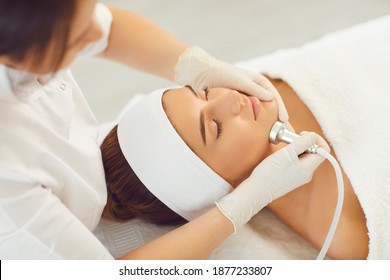 Cosmetologist making procedure of microdermabrasion of facial skin for young serene woman patient in neauty salon, top view. Cosmetology and professional skin care concept