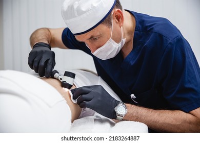 A cosmetologist male doctor in black gloves and blue uniform examines the patient's mole using a dermatoscope. 