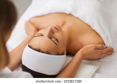 Cosmetologist or masseur making manual relaxing rejuvenating massage for face and shoulders for young woman in beauty salon, rear view. Rejuvenating facial massage in cosmetology