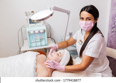 A cosmetologist in a medical mask and rubber gloves applies a cream mask to the client's face. Top view. In the background, a cosmetology device. Concept of cosmetology during the pandemic