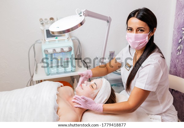 A cosmetologist in a medical mask and rubber\
gloves applies a cream mask to the client\'s face. Top view. In the\
background, a cosmetology device. Concept of cosmetology during the\
pandemic